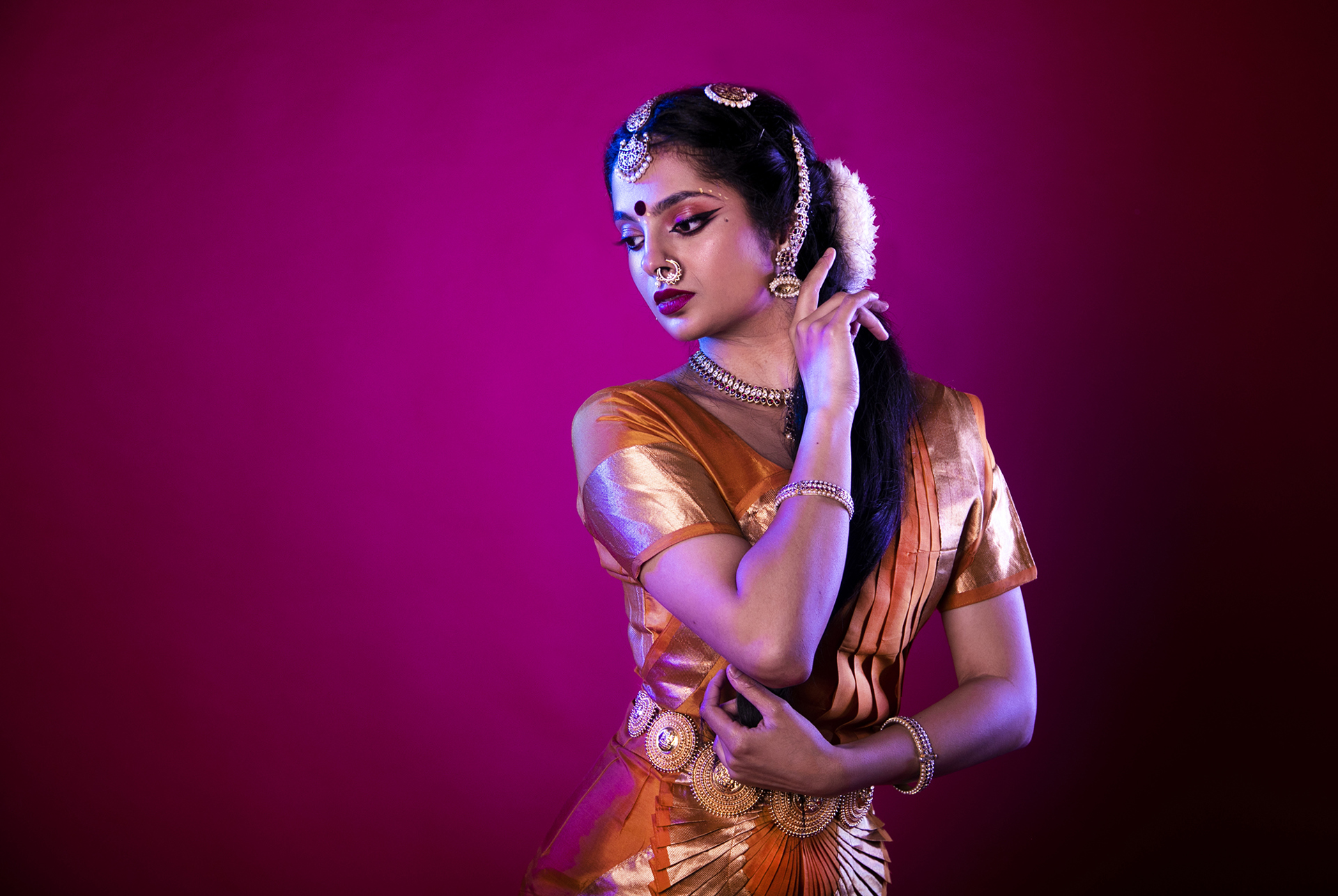 South Asian dancer holding one hand to face and the other towards her elbow on hot pink studio backdrop. Wearing traditional orange dress with gold belt bracelets necklace and head jewlery with gold nose ring.