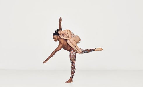 Global majority male dancer on one leg with arms stretched wise with global majority female dancer curled up balancing in his back in a white studio