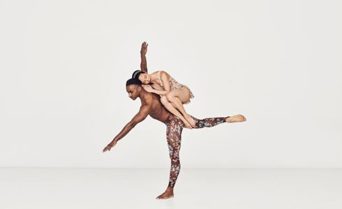 Global majority male dancer on one leg with arms stretched wise with global majority female dancer curled up balancing in his back in a white studio