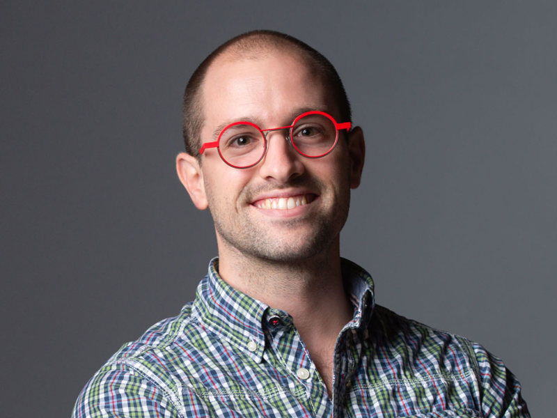 Headshot of Constant Vigier. White male with shaved head smiling at camera wearing red circle glasses and checked shirt 