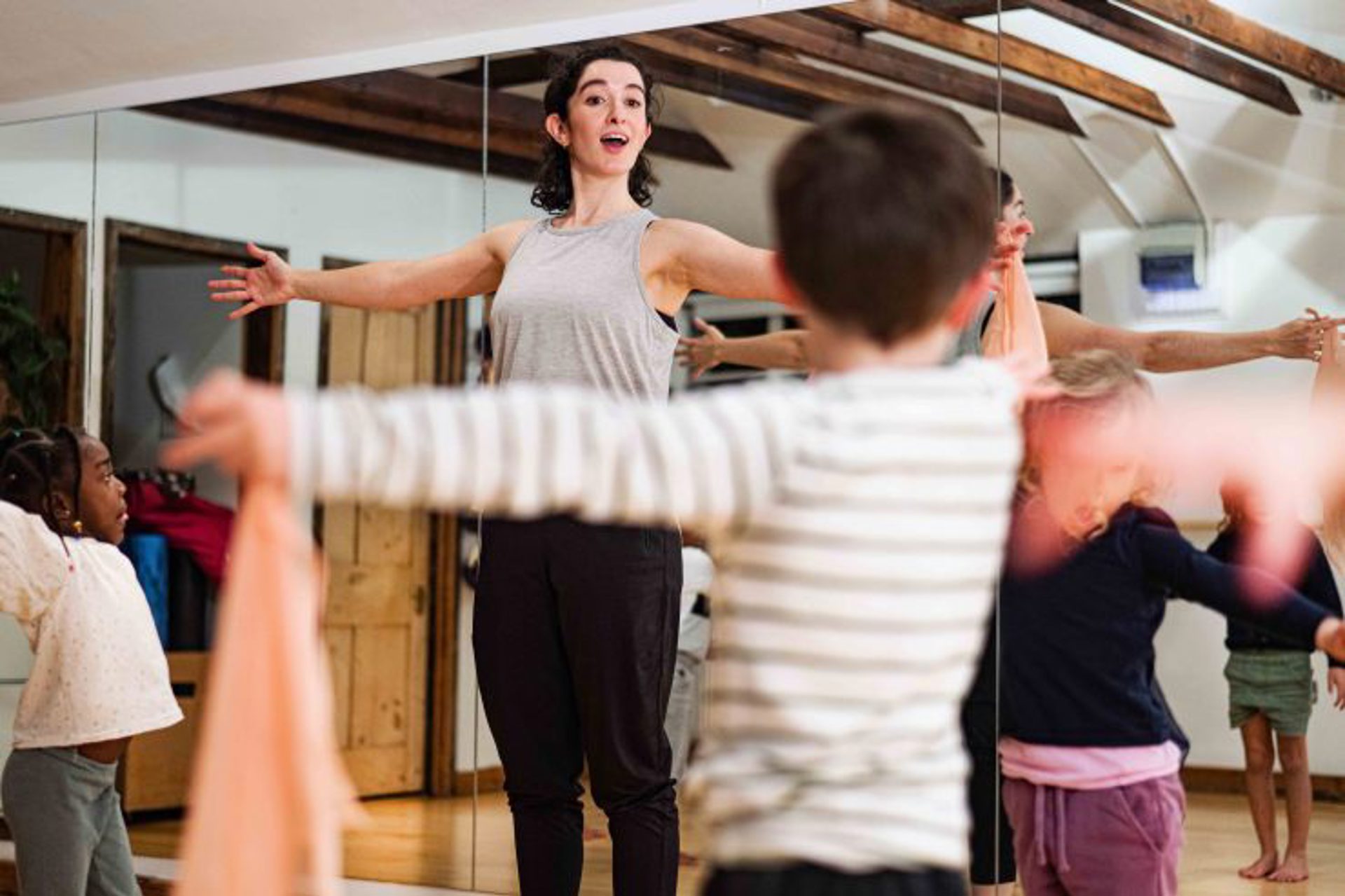 White female dancer with arms stretched wide teaching a class of small children in a dance studio with mirrored walls 