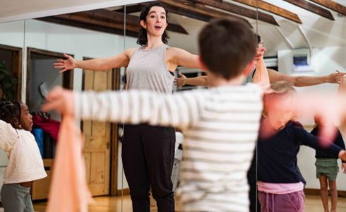 White female dancer with arms stretched wide teaching a class of small children in a dance studio with mirrored walls 