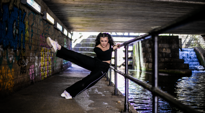 white female dancer with brown curly hair leaning on a raining undernieth a tunnel with water to the right side of the image. wearing blacktracksuit and white trainers