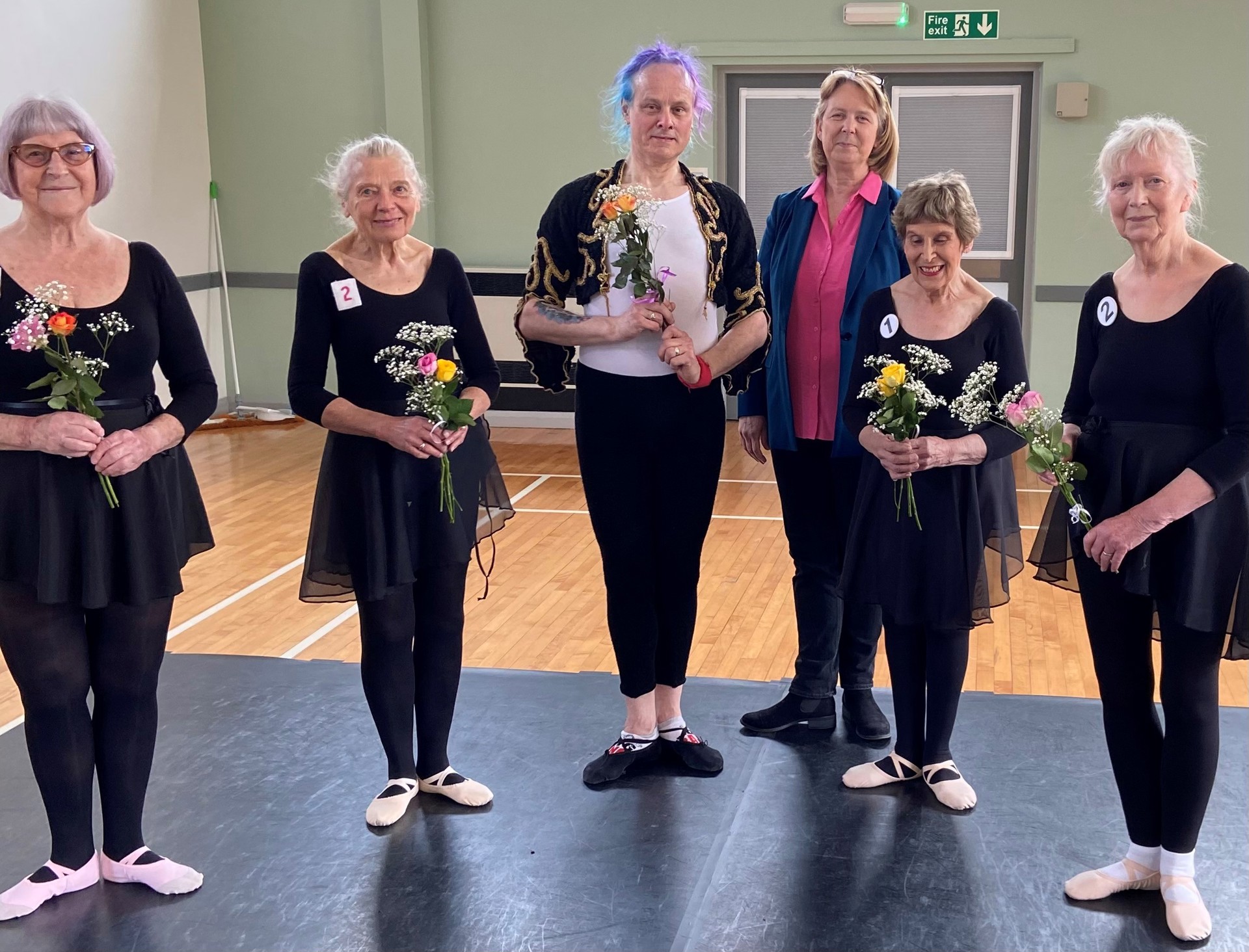 Six older white Bbodance dancers wearing black ballet costumes holding flowers and looking at the camera. In a dance studio,