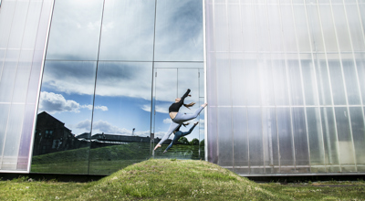 female dancer jumping with one leg pointed towards the ground and the other kicked behind witharms in the air on a grassey hill. In front of metal Trinity Laban University building  with mirroed wall reflecting clouded sky. Wearing grey leggings and black crop top. 