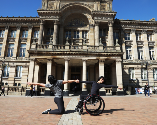 White brunette female dancer wearing all black in a wheel chair holding hands with a white brunette femle dancer on her knees both stretching out the the other arm away from each other. Outside a large pillared building. 