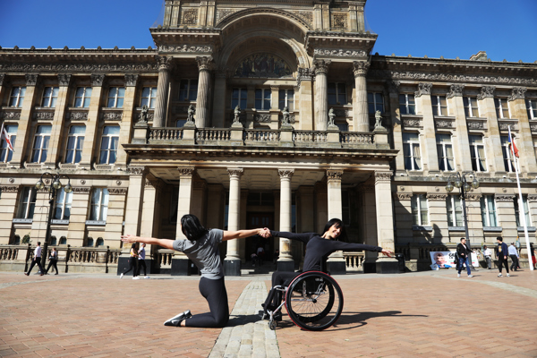 White brunette female dancer wearing all black in a wheel chair holding hands with a white brunette femle dancer on her knees both stretching out the the other arm away from each other. Outside a large pillared building. 