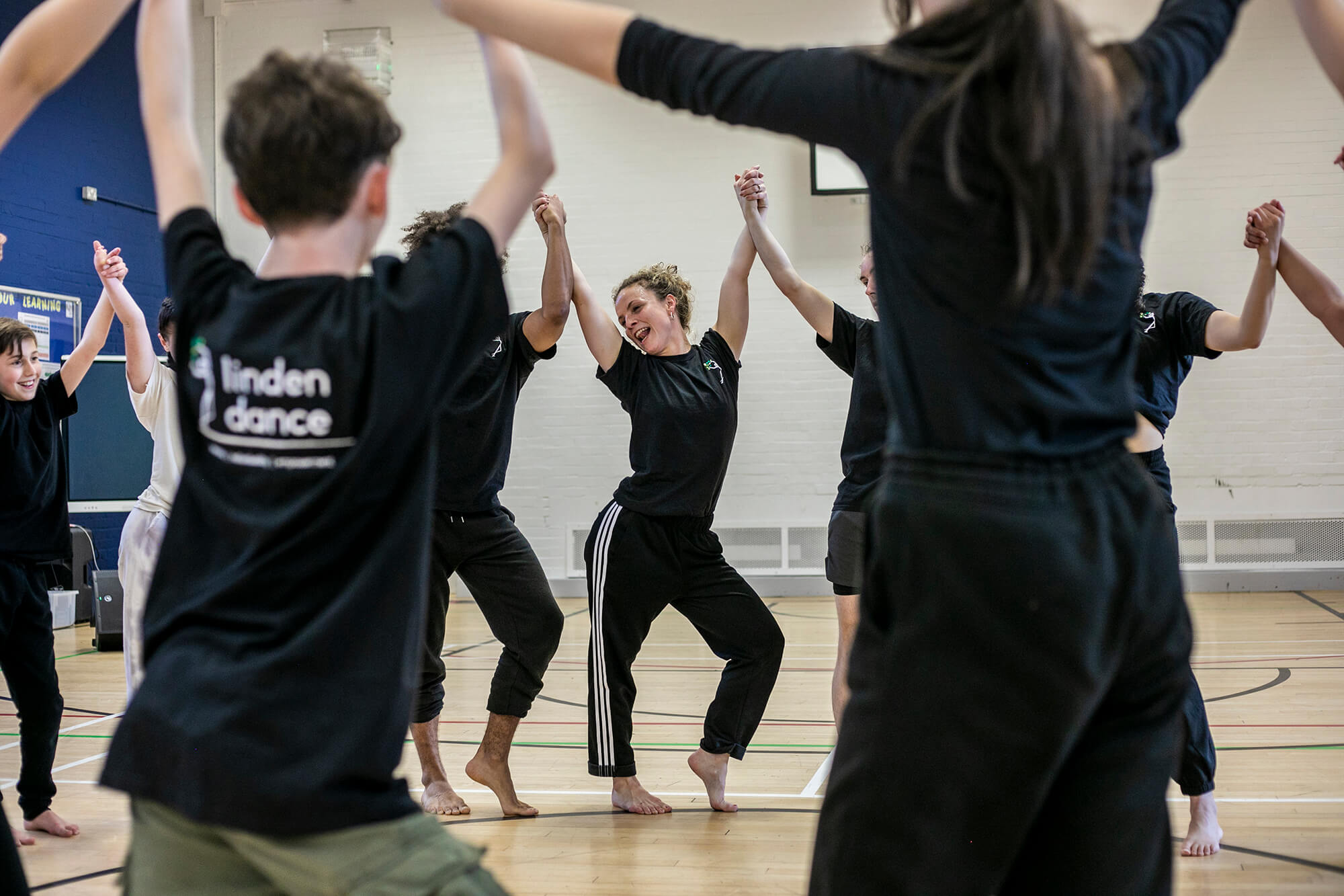 Dance circle with young people holding hands with the joint hands in the air. ca,era focused on the teacher with hip popped to the side and smiling. All wearing black PE kit in a sports hall.