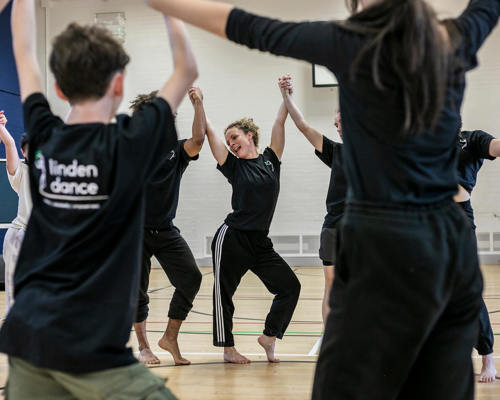 Dance circle with young people holding hands with the joint hands in the air. ca,era focused on the teacher with hip popped to the side and smiling. All wearing black PE kit in a sports hall.