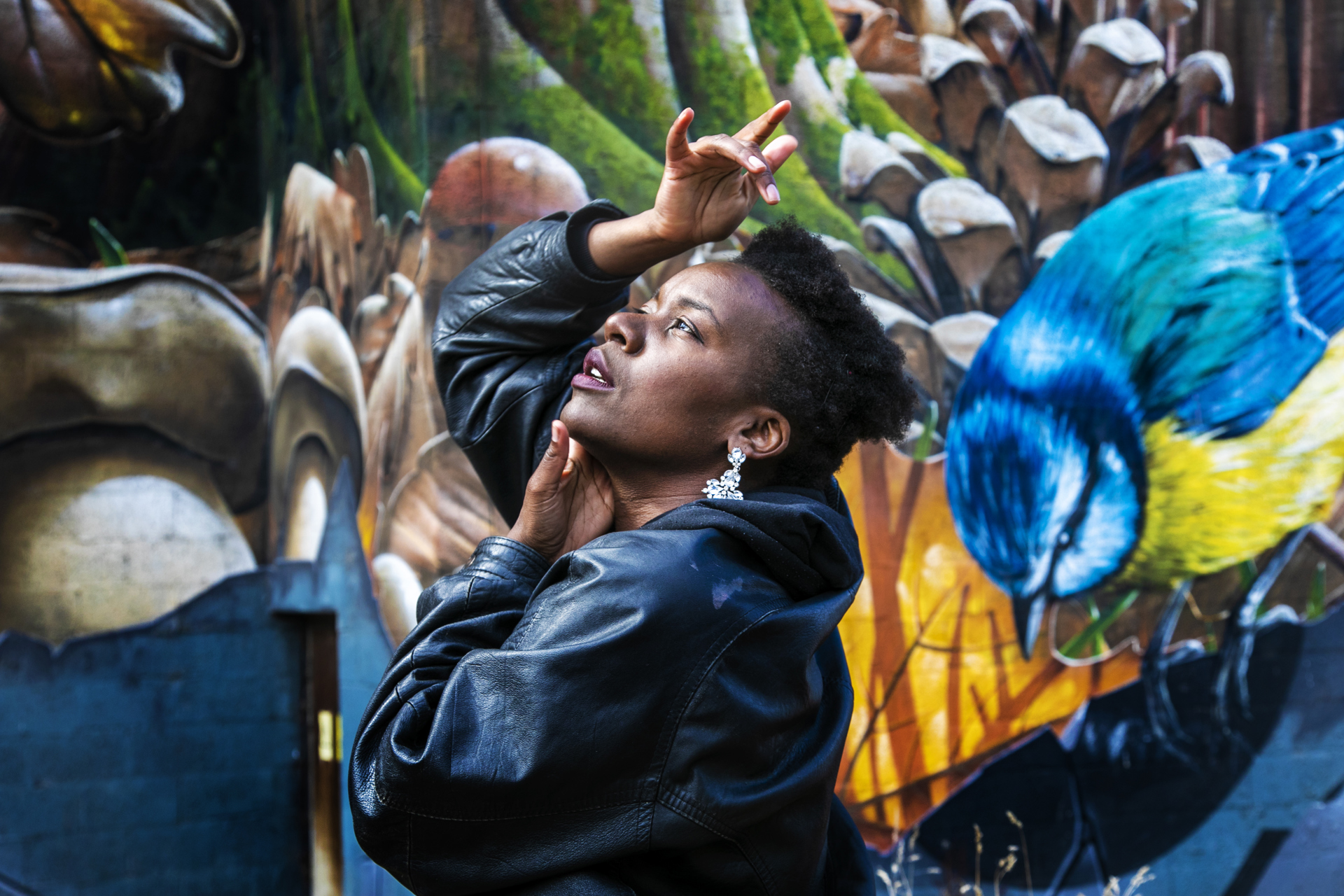 Global Majority female looking up to the top left of the image with hands around her face. Wearing a black leather jacket with short afro hair. In front of colourful graffiti wall