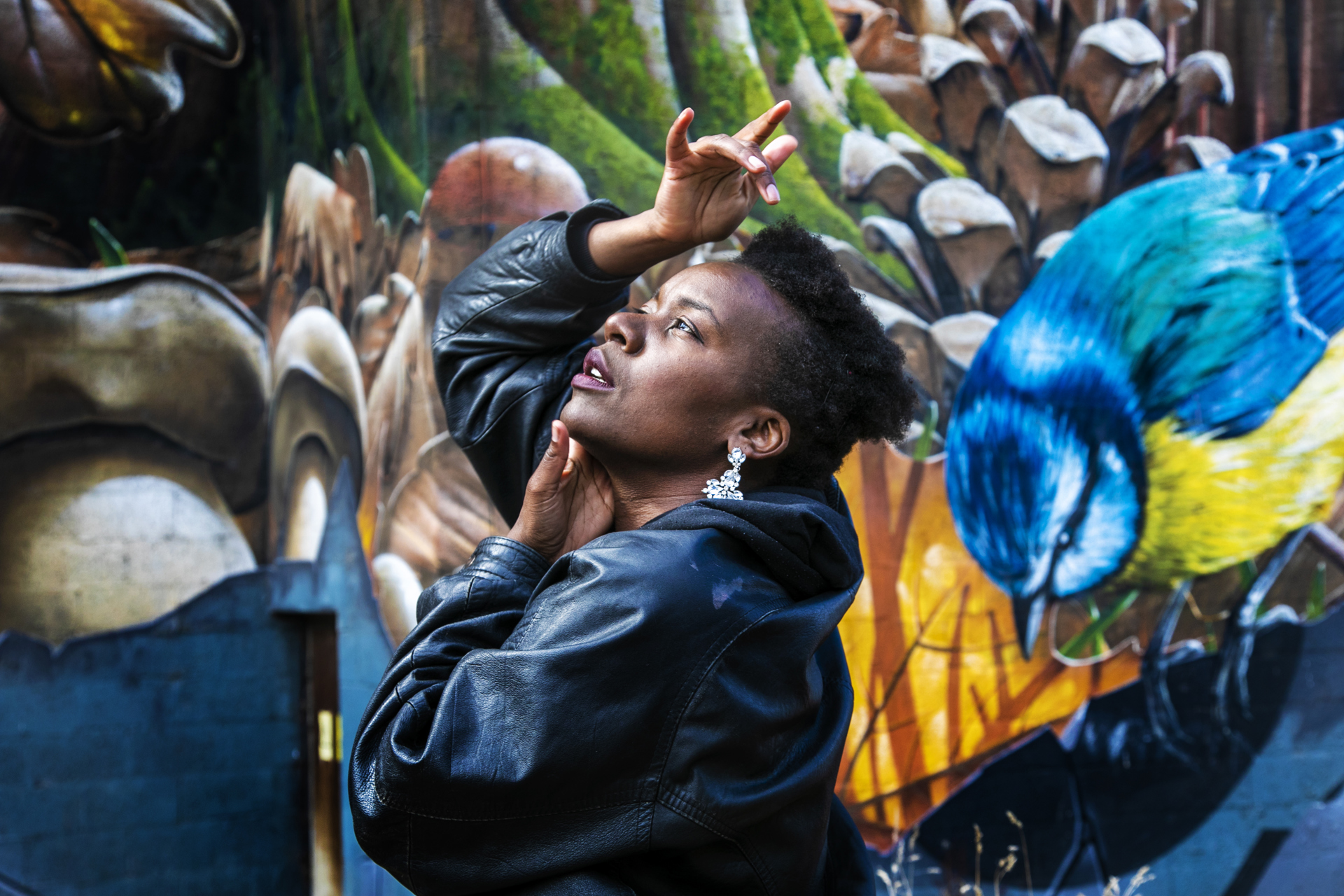Global Majority female looking up to the top left of the image with hands around her face. Wearing a black leather jacket with short afro hair. In front of colourful graffiti wall Global Majority female looking up to the top left of the image with hands around her face. Wearing a black leather jacket with short afro hair. In front of colourful graffiti wall