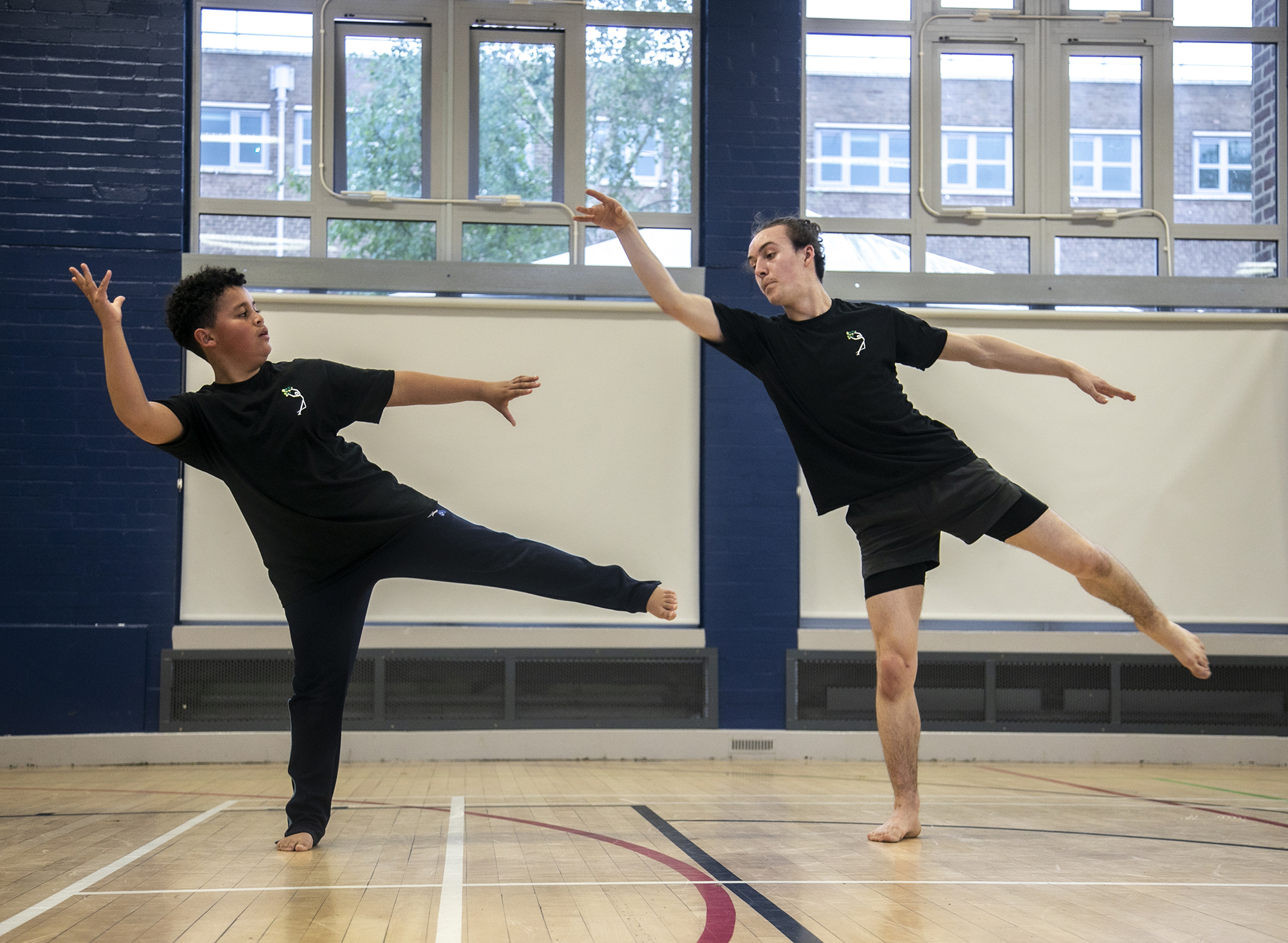 teenage male dance teacher on one leg reaching to the top left of the image teaching young boy dancer who is mirroring the arms and the legs. In sports hall both wearing black PE kit teenage male dance teacher on one leg reaching to the top left of the image teaching young boy dancer who is mirroring the arms and the legs. In sports hall both wearing black PE kit
