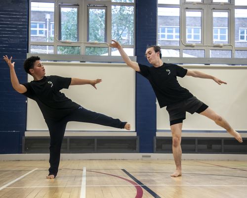 teenage male dance teacher on one leg reaching to the top left of the image teaching young boy dancer who is mirroring the arms and the legs. In sports hall both wearing black PE kit