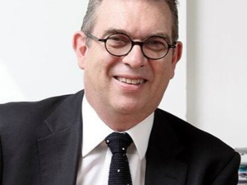 Headshot Anthony Bowne. White male smiling with circular glasses. Short grey hair, wearing a black suit, white shirt, and black tie.