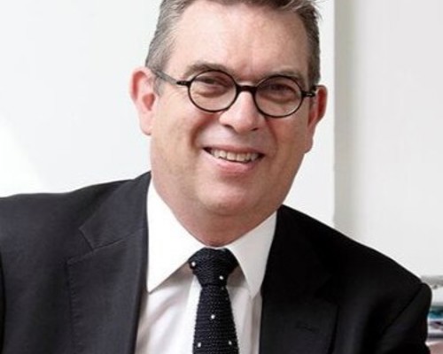 Headshot Anthony Bowne. White male smiling with circular glasses. Short grey hair, wearing a black suit, white shirt, and black tie.