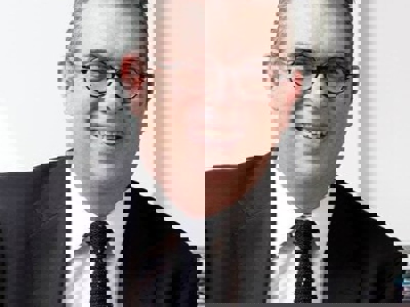 Headshot Anthony Bowne. White male smiling with circular glasses. Short grey hair, wearing a black suit, white shirt, and black tie. Headshot Anthony Bowne. White male smiling with circular glasses. Short grey hair, wearing a black suit, white shirt, and black tie.