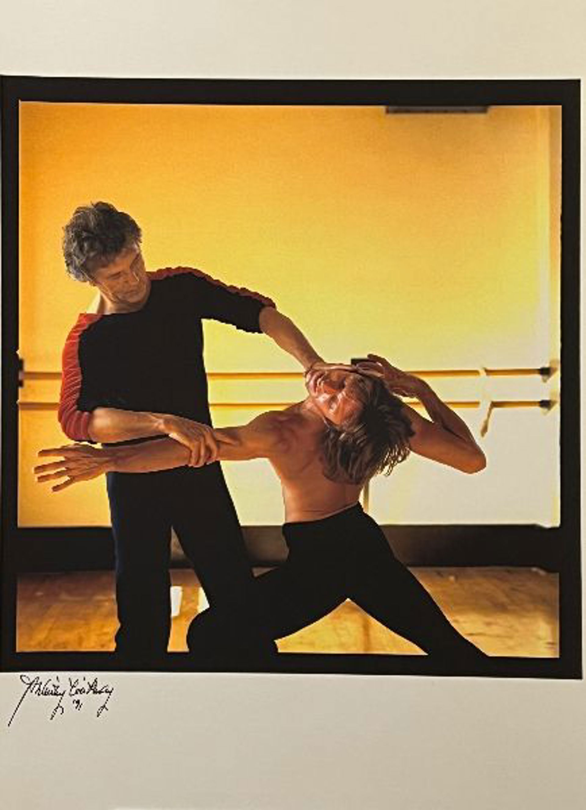 male dance teacher holding male dancers arm and head in movement in front of ballet bar in an orange lit dance studio