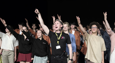 White male dance teacher standing in the centre of a stage with one hand raised to the top left looking in the same direction with a large group of young people copying the movement behind. 