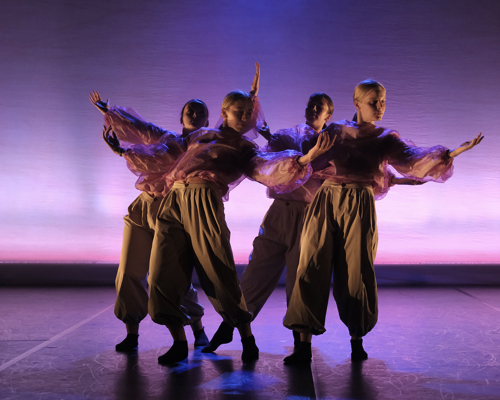 4 female dancers wearing baggy pink tops and cargo trousers all huddled together with arms stretched out leaning towards eachother. Stage lighting is pink and purple