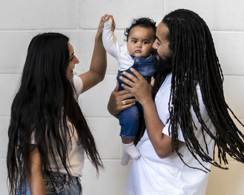 Family of 3. Global majority male with long dreadlocked hair holding baby, global majority female with long black hair, holding the babies hand as they dance. Wearing blue jeans, trainers and white tshirts infront of white brick wall. 