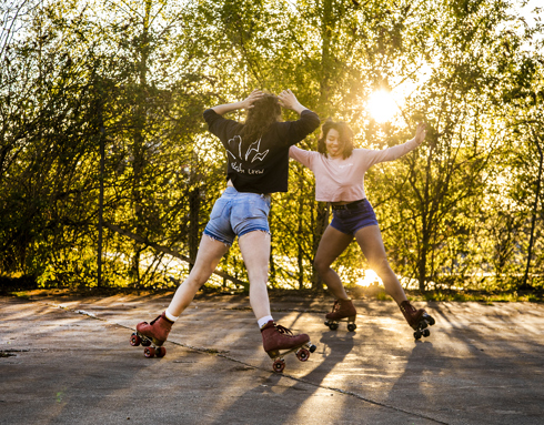 two female roller skaters skating around each other with smiles in golden dappled light with trees in the background 