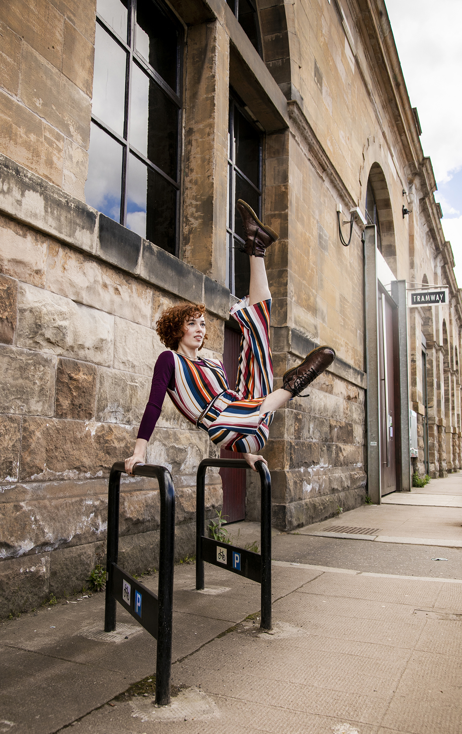 white female dancer with red short curly hair and striped jumpsuit, swinging on two metal bike stands high kicking both legs in the air. In front tramway brick building on the street.