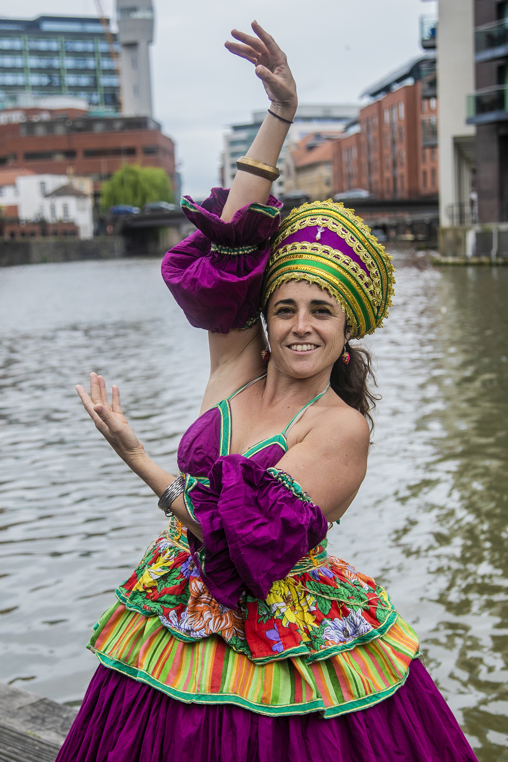 Brazialian female dancer brown hair wearing traditional purple and and gold dress with large golden headpiece holding arm above her head and one framing the face. In front of river and urban Bristol buildings.