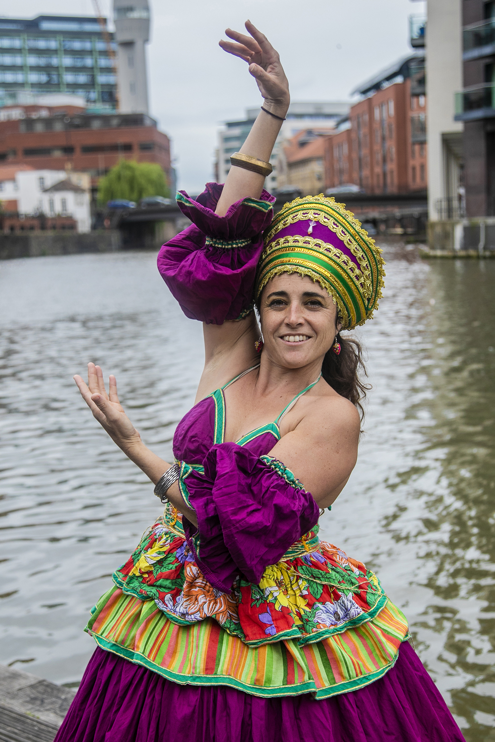 Brazialian female dancer brown hair wearing traditional purple and and gold dress with large golden headpiece holding arm above her head and one framing the face. In front of river and urban Bristol buildings. Brazialian female dancer brown hair wearing traditional purple and and gold dress with large golden headpiece holding arm above her head and one framing the face. In front of river and urban Bristol buildings.