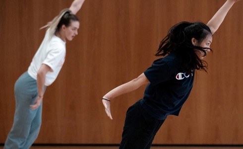 two young female dancers leaning diagonally with one arm up and other by their side. In front of wooden wall