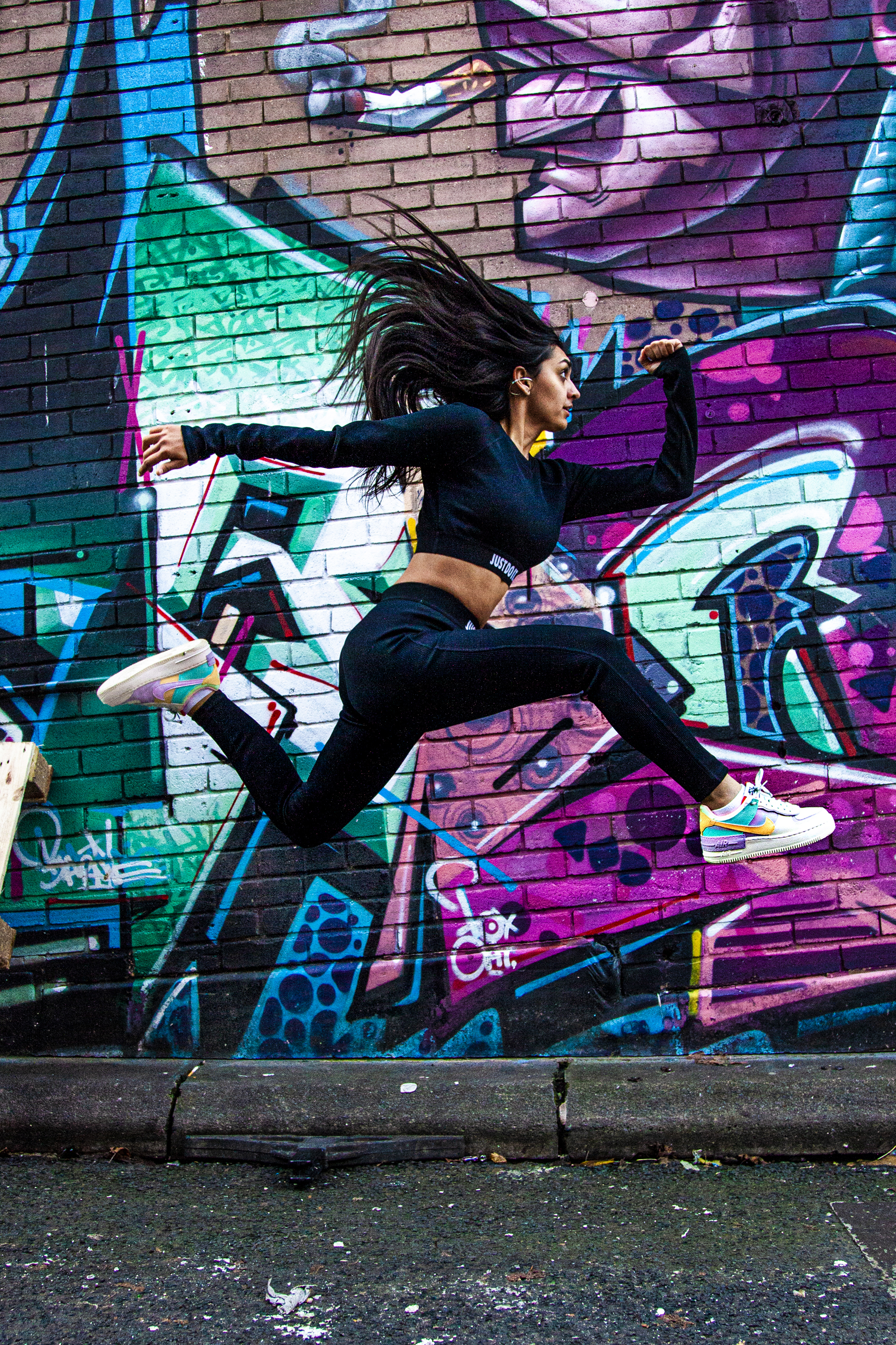 Global majority female dancer jumping in a running pose side on with long black hair swishing behind. Wearing black leggings and long sleeve black top in front of purple graffiti wall
