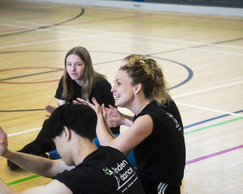 white female teacher with teenage student sitting on a sports hall floor with hands gesturing. All wearing black with linden dance logo on the t-shirts. 