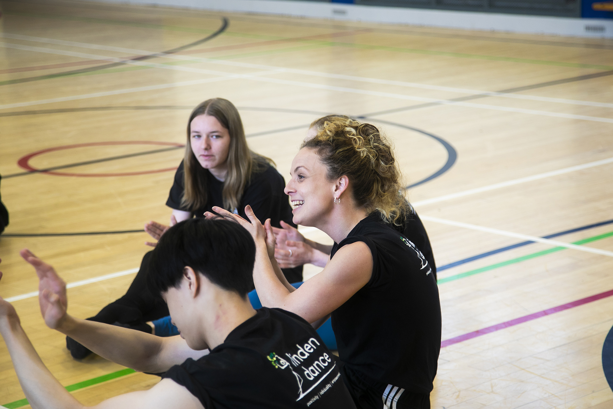 white female teacher with teenage student sitting on a sports hall floor with hands gesturing. All wearing black with linden dance logo on the t-shirts. 
