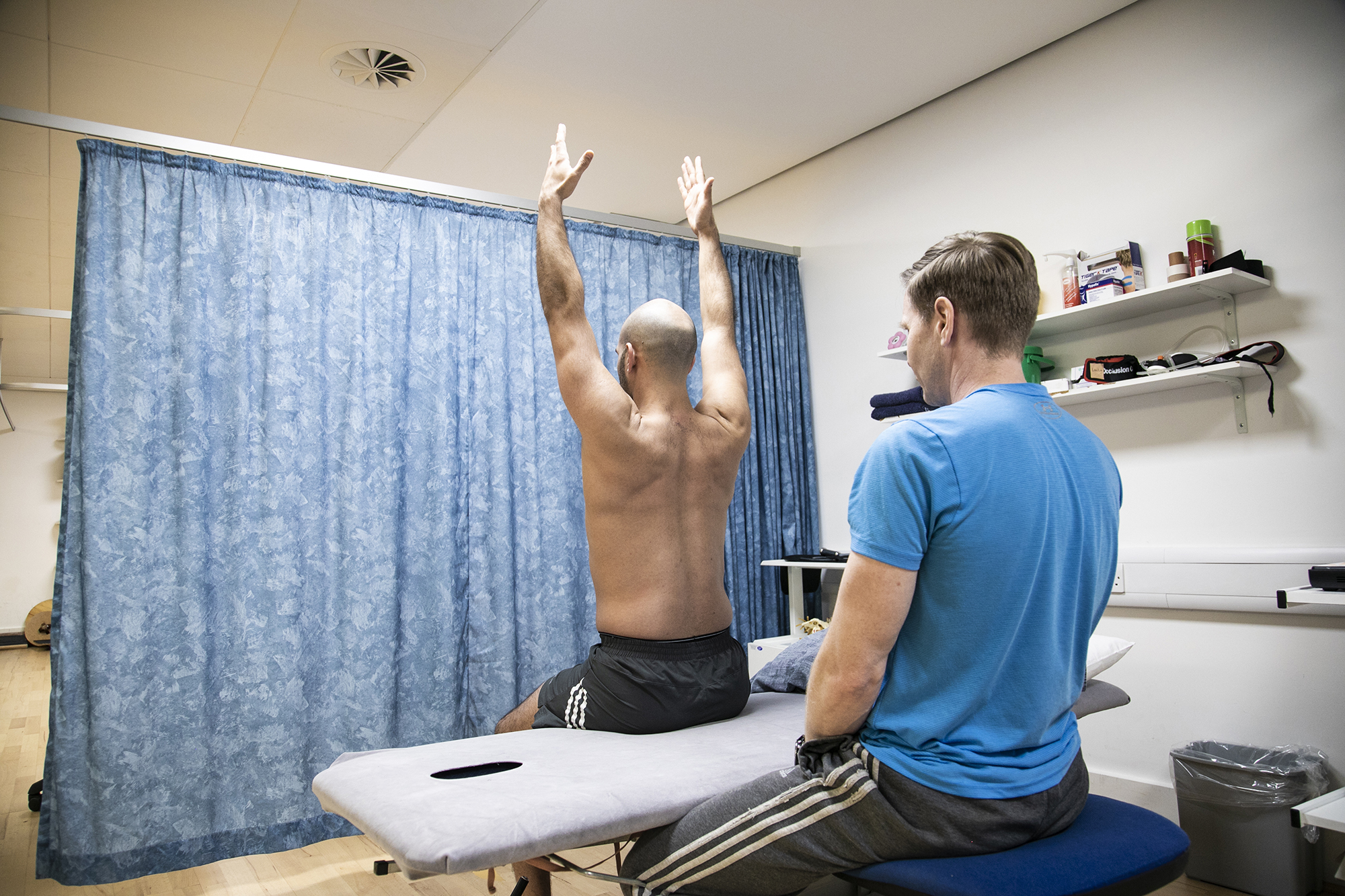 Male dancer sitting on physio bed with back to camera and arms up in the air, with male physiotherapist looking at dancer in an examination. With blue curtain in the background and a shelf of physio equipment. 
