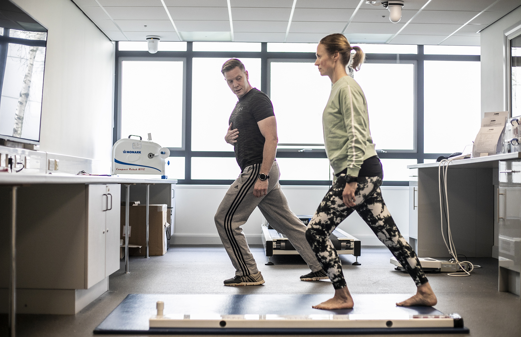 white male physio lunging forward holding his stomach, with white female dancer mirroring his movement. Both wearing excersise clothes in a bright healthcare labratory with large windows in the background.