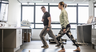 white male physio lunging forward holding his stomach, with white female dancer mirroring his movement. Both wearing excersise clothes in a bright healthcare labratory with large windows in the background.