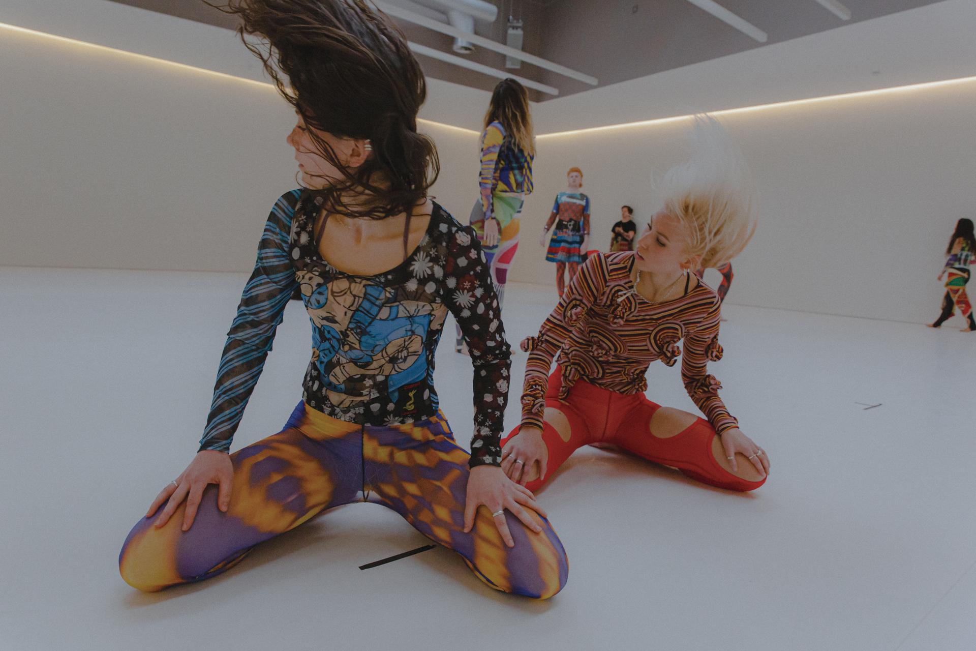 two female dancers sitting on their knees with hands on knees looking left and flicking hair. Wearing colourful patterned clothes 