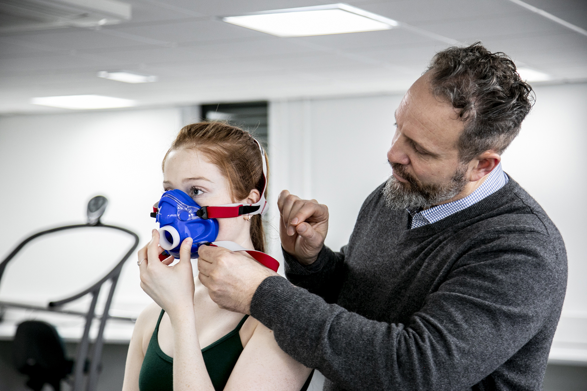 White female dancer with long red hair with white male physiologisttightening face-mask to the dancer to measure cardiovascular fitness. In a white laboratory setting.