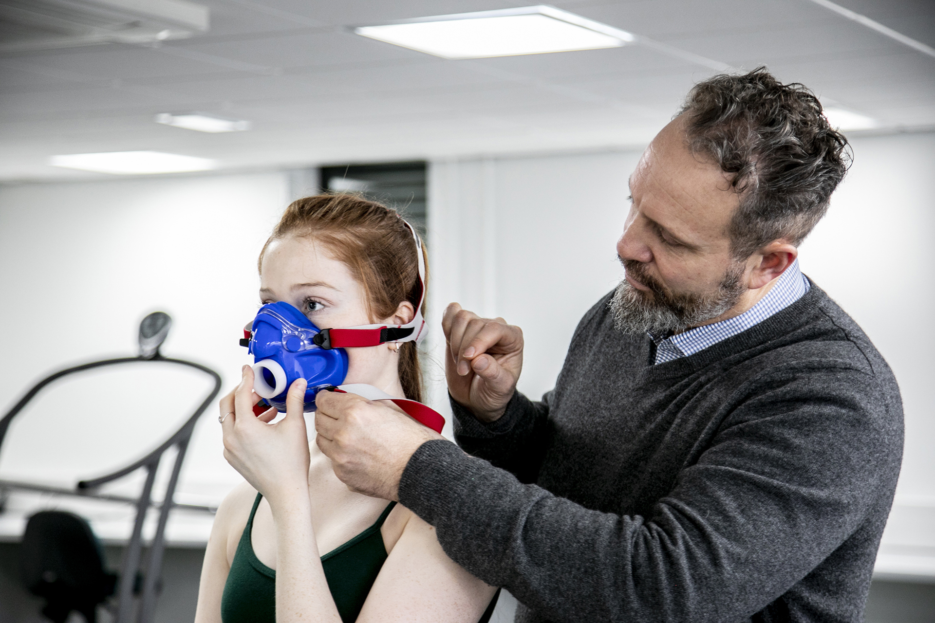 White female dancer with long red hair with white male physiologisttightening face-mask to the dancer to measure cardiovascular fitness. In a white laboratory setting. White female dancer with long red hair with white male physiologisttightening face-mask to the dancer to measure cardiovascular fitness. In a white laboratory setting.