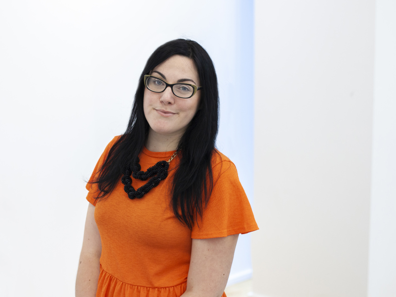 Headshot of Lara Coffey white female with black long hair and glasses smiling at the camera. Wearing orange top 