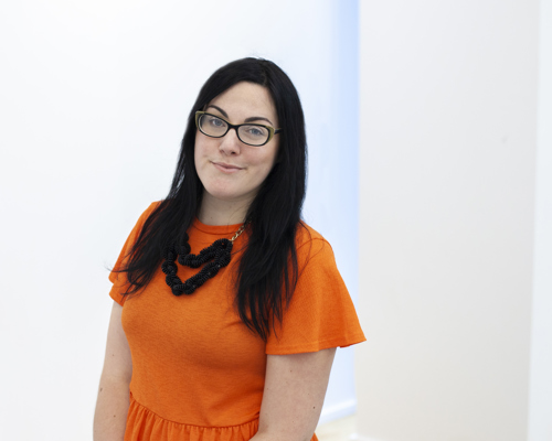 Headshot of Lara Coffey white female with black long hair and glasses smiling at the camera. Wearing orange top 