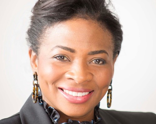 Headshot of Denise Nurse. Global majority female with tied back dark hair, smiling. Wearing large hoop earrings, black blazer and ruffled black top. 
