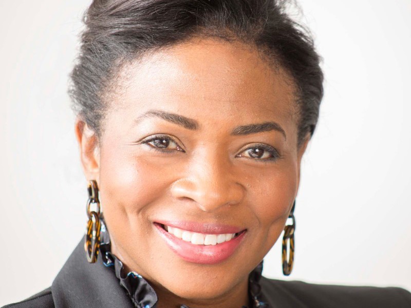 Headshot of Denise Nurse. Global majority female with tied back dark hair, smiling. Wearing large hoop earrings, black blazer and ruffled black top. 