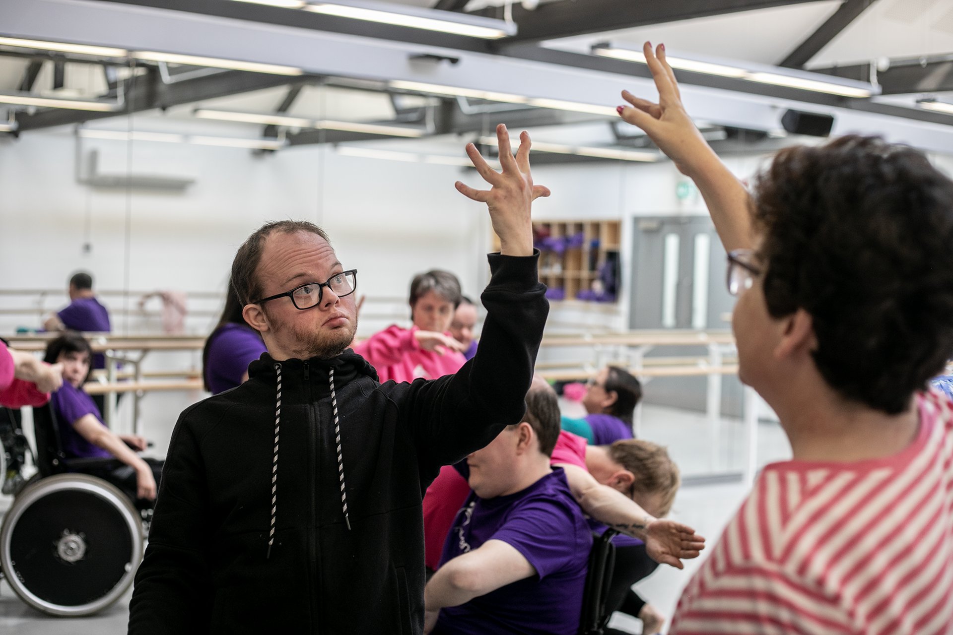 community dance group in the dance studio. Main focus on male downs syndrome dancer with hand stretched wide with other another dancer mirroring it. 