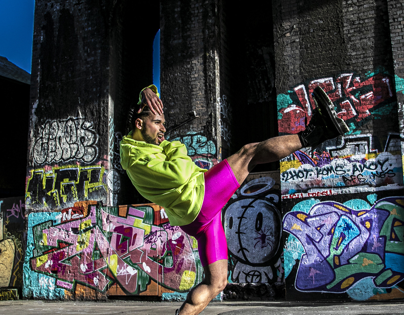 LGBTQ+ global majority male dancer with short dreads wearing neon yellow jumper with pink leotard shorts and black boots. Hands around the face, body leaning back standing on one tip toe with one leg kicked out. In front of colourful graffiti wall.