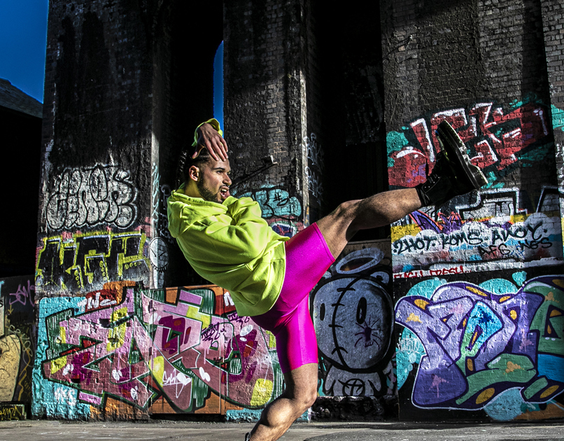 LGBTQ+ global majority male dancer with short dreads wearing neon yellow jumper with pink leotard shorts and black boots. Hands around the face, body leaning back standing on one tip toe with one leg kicked out. In front of colourful graffiti wall. LGBTQ+ global majority male dancer with short dreads wearing neon yellow jumper with pink leotard shorts and black boots. Hands around the face, body leaning back standing on one tip toe with one leg kicked out. In front of colourful graffiti wall.