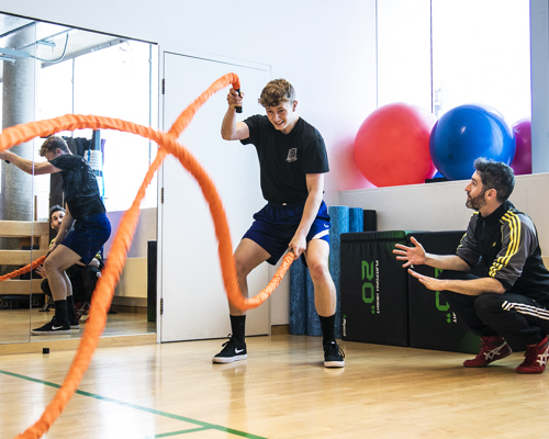 Two white males in a gym, one slamming down orange ropes the other male squatting down next to him. With a mirrored wall and exercise equipment in the background. 📄 Screenshot 2023-07-06 at 16.05.38.png   Two white males in a gym, one exercising with a large orange battle rope while the other male squats down beside him. There is a mirrored wall and exercise equipment in the background.