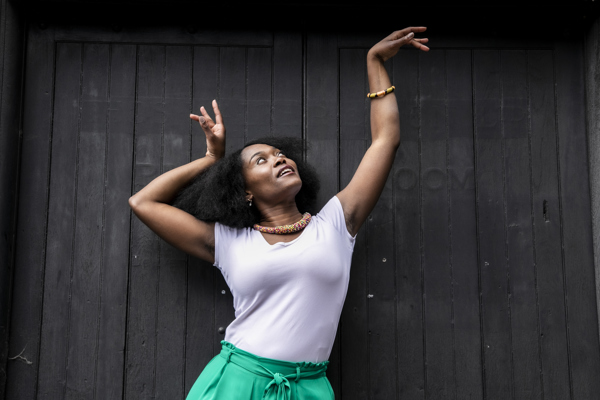 Global majority female dancer with afro, holding one arm out to the top right of the image with eyes looking at a limp wrist and the other hand next to her head. Wearing white top, green trousers and colourful beaded necklace in fornt of black wall