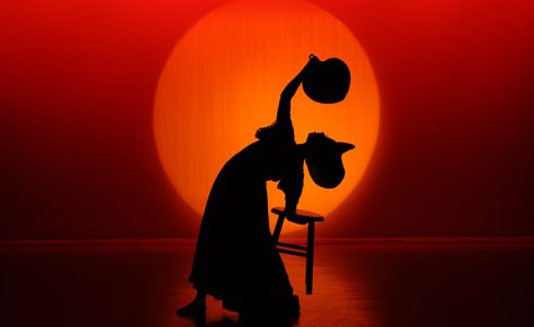 silhouette of dancer doing a backbend holding a tool with one hand and arching over waving a fan to the face wearing a large hat. In front of large orange circle and red background