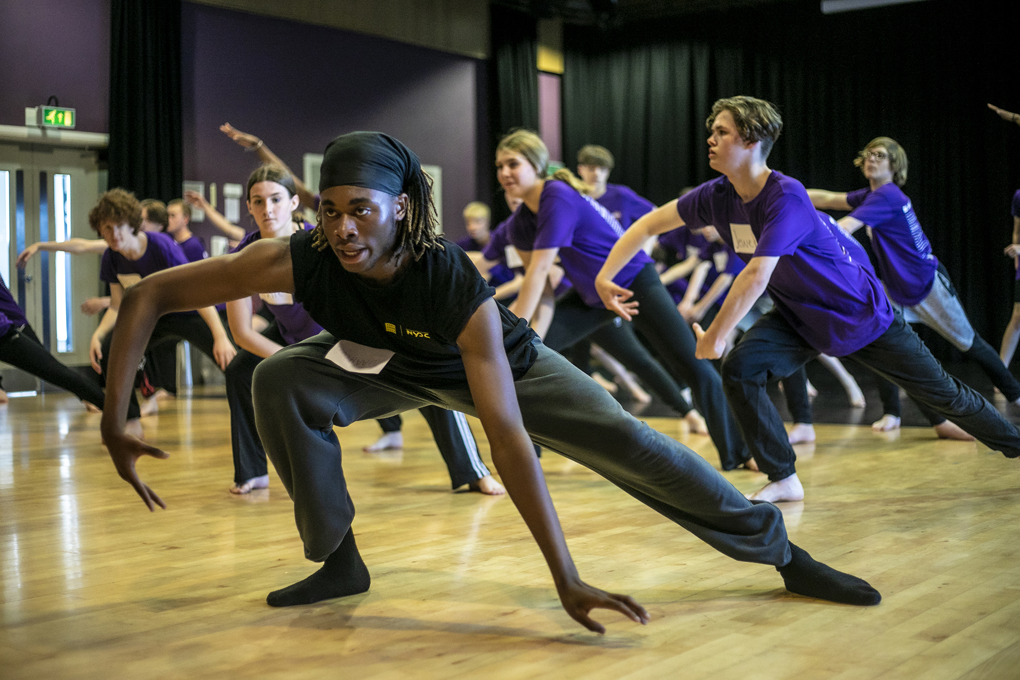 black male dance artist teaching room of young people, teacher and students in deep lunges with one hand touching the floor the other in a wing shape. Teacher wearing black students out of focus wearing purple t-shirts. 