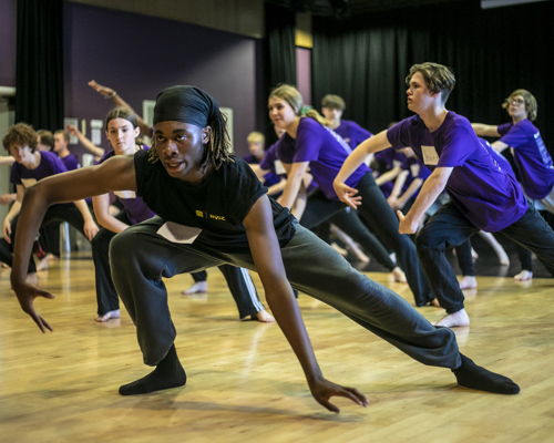 black male dance artist teaching room of young people, teacher and students in deep lunges with one hand touching the floor the other in a wing shape. Teacher wearing black students out of focus wearing purple t-shirts. 
